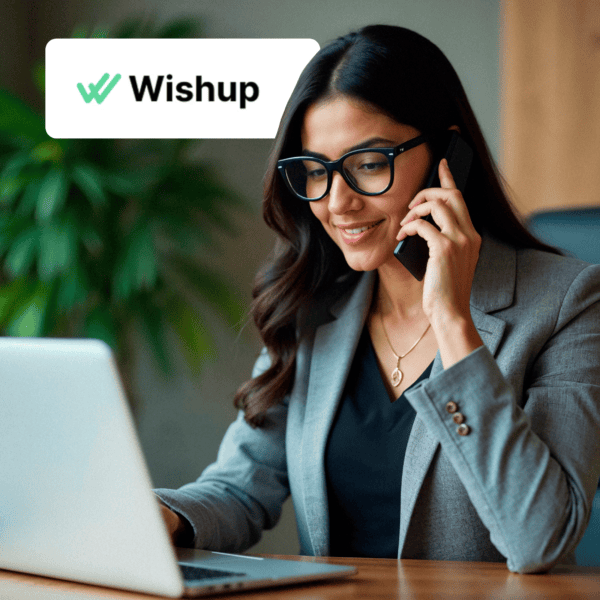 A professional woman smiling while talking on her phone and looking at a laptop, with the Wishup logo in the top left corner.